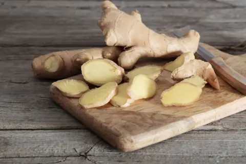 Frischer Ingwer, teils in Scheiben geschnitten, liegt auf einem Holzbrett. Der Unterschied zwischen ganzen Wurzeln und den Scheiben ist deutlich zu erkennen.