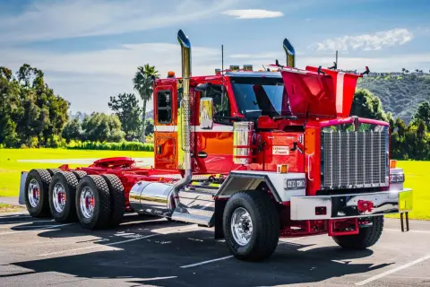 Un impressionnant Kenworth, un camion américain rouge vif, brille sous le soleil. Ses chromes étincelants et ses échappements imposants annoncent sa puissance.