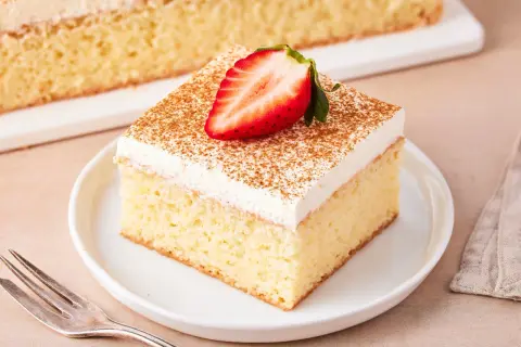 Ein Stück saftiger Tres Leches Kuchen, garniert mit einer Erdbeerhälfte und Zimt, auf einem Teller mit einer Gabel daneben.
