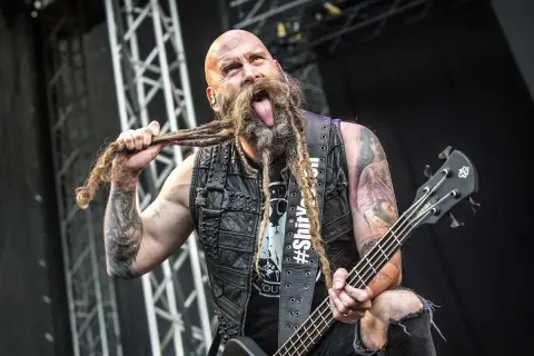 Chris Kael, basista z długą, zaplecioną brodą, wyciąga język podczas koncertu, trzymając gitarę basową.