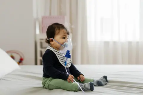 Małe dziecko w maseczce inhalacyjnej. Delikatna para unosząca się z nebulizatora świadczy o tym, że odbywają się inhalacje dla niemowlaka.