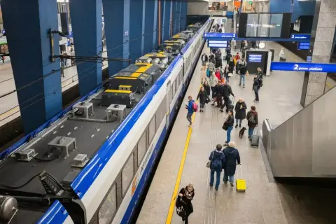 Ludzie z bagażem czekają na peronie PKP. Widać pociąg, tablice informacyjne i schody ruchome.