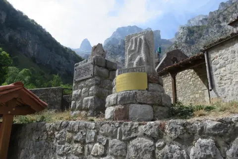Monumento en piedra con inscripciones y un relieve, con montañas al fondo. El cartel dice "EL CAIN DE ARRIBA".