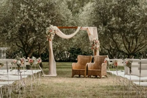 Un décor champêtre pour une célébration laïque : chaises blanches, arche fleurie, fauteuils en rotin sous les oliviers.