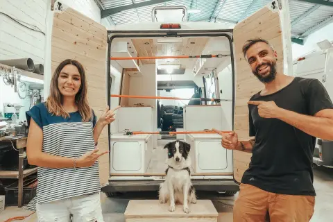 Pareja sonriente y su perro posan junto a una furgoneta camper en proceso de construcción, ¡las mejores furgonetas camper calidad precio!