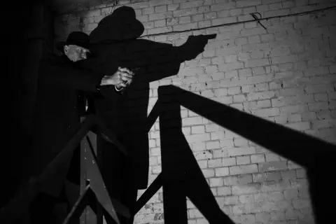 A man in a fedora and trench coat aims a pistol in this dramatic film noir photography. His shadow looms large against a brick wall.