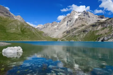 Le lac de la Sassière, aux eaux turquoise, reflète les montagnes majestueuses et le ciel bleu. Un rocher émerge, témoin de la quiétude du paysage.