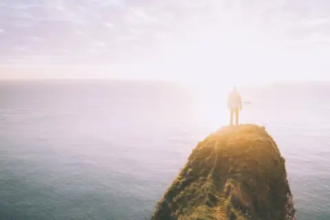 Ein Mensch steht auf einer Klippe am Meer, die Sonne scheint hell. Ein Symbol für das Durchhalten in schweren Zeiten, wenn man dem Horizont entgegenblickt.