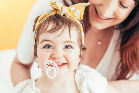A baby with bright blue eyes and a yellow headband smiles, her passy teeth peeking out from behind a pacifier. Her mother smiles in the background.