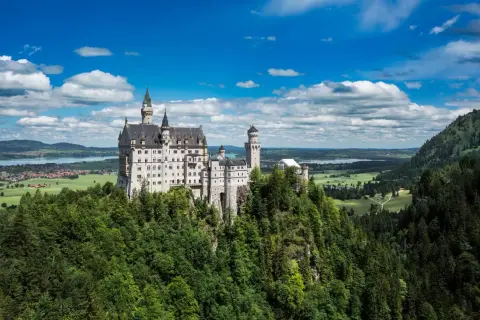El castillo del rey loco, Neuschwanstein, se alza majestuoso sobre un risco verde, con torres que acarician las nubes y un paisaje de ensueño.