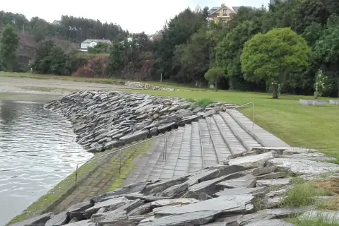 Escaleras de piedra y césped verde junto al agua en la playa de los bloques Ribadeo, con casas y árboles al fondo.