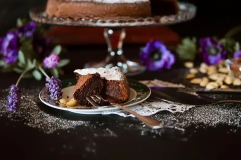 Ein Stück saftiger italienischer Schokokuchen, bestäubt mit Puderzucker, auf einem Teller mit Mandeln und lila Blumen.