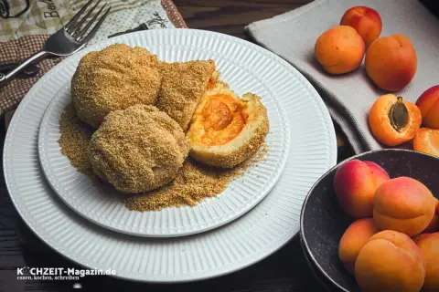 Mehlspeisen aus Kartoffelteig mit Aprikosenfüllung, paniert und auf einem Teller mit zerbröselten Semmelbröseln angerichtet. Daneben frische Aprikosen.