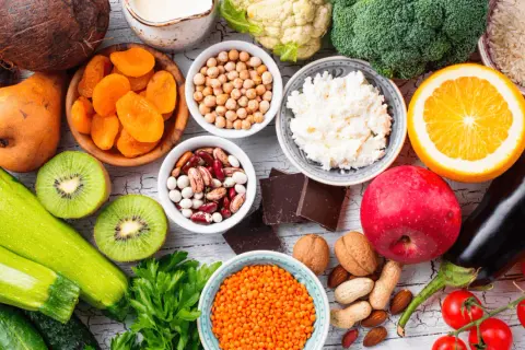 Kolorowy stół pełen zdrowych produktów: cukinia, kiwi, pomidory, bakłażan, jabłko, pomarańcza, brokuły, kalafior, soczewica, ciecierzyca, fasola, orzechy, migdały, suszone morele, ryż, twaróg i gorzka czekolada – idealne warzywa o niskim indeksie glike...