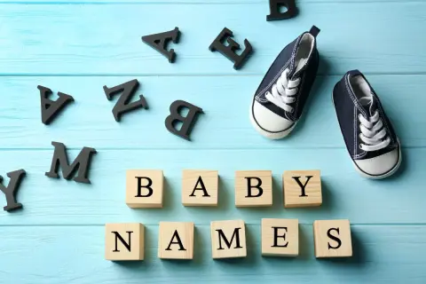 Des lettres noires forment "BABY NAMES" sur des cubes en bois. Une paire de chaussons bleus attend. Quel joli prénom en M pour le bébé ?