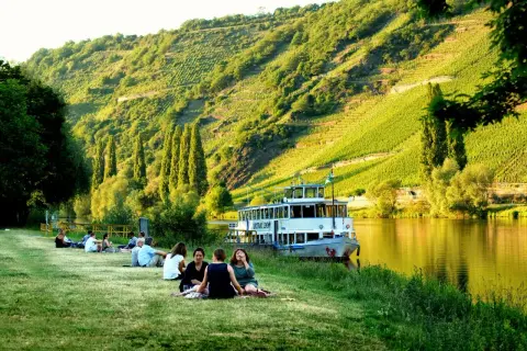 Entspannte Menschen genießen die Sonne am Ufer der Mosel. Ein Ausflugsschiff liegt vor, im Hintergrund steile Weinberge. Perfekte Mosel Ausflugsziele.