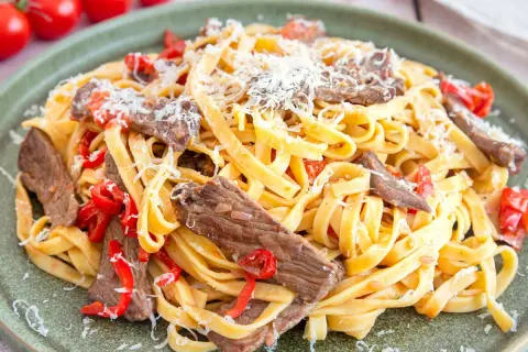 Herzhaftes Gericht mit Pasta und zarten Rinderfiletstreifen, garniert mit geriebenem Parmesan und roten Paprikastücken.