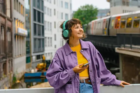 Junge Frau mit Kopfhörern und Handy in der Hand, die auf eine Stadtkulisse mit Zug blickt. Sie plant ihr Fachhochschulreife Fernstudium.