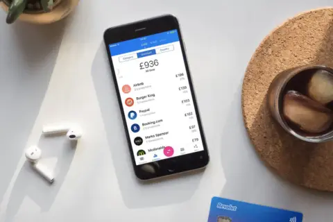 Telefon z aplikacją Revolut, pokazujący wydatki i opcje, np. revolut ubezpieczenie podróżne. Obok słuchawki i drink.