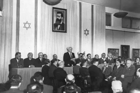 Hombres en traje asisten a un evento histórico, la creacion del estado de Israel, con la bandera israelí y un retrato de Theodor Herzl.