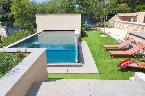 Un magnifique bassin vitré aux eaux bleues, entouré d'une pelouse synthétique et de chaises longues roses.