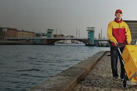 Kurier DHL z wózkiem i paczką do Włoch na tle kanału i miasta.