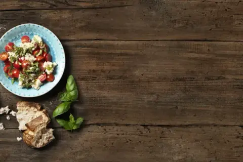 Två tallrikar med tomatsallad och vispad mozzarella, garnerad med basilika. Körsbärstomater och bröd på träbord.