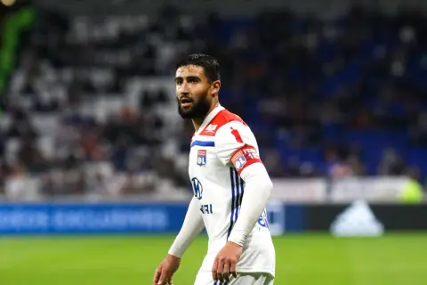 Piłkarz Nabil Fekir w białej koszulce z czerwonymi elementami, z opaską kapitańską na ramieniu, na tle trybun.