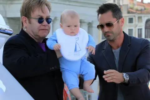 Elton John z dziećmi, w tym małym dzieckiem w niebieskim ubranku i śliniaczku z łódką, w otoczeniu innych mężczyzn.
