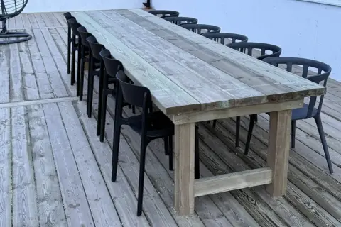 Ett stadigt, hemmabyggt utebord av trä med plats för många gäster. Svarta stolar står uppradade runt det robusta bordet på altanen.
