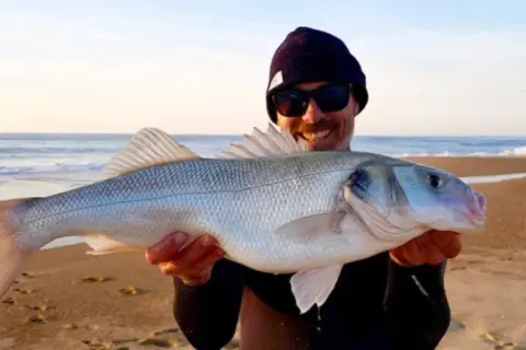Glücklicher Angler hält stolz einen schönen Barsch Salzwasser am Strand.