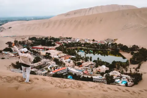 Oaza na pustyni Huacachina, z turystką na wydmie pozdrawiającą zieloną, bujną okolicę z jeziorem i hotelami.