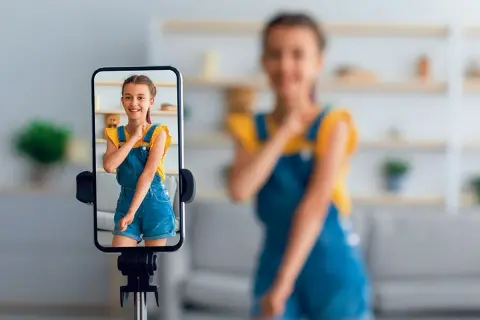 Dziewczynka na ekranie telefonu uśmiecha się, podczas gdy mama w tle nagrywa filmik. To przykład zjawiska sharenting co to znaczy dzielenie się życiem dzieci w sieci.
