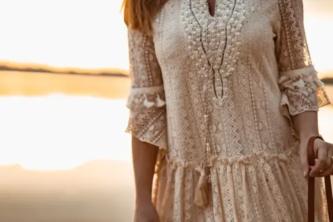 Kobieta w koronkowej sukience w stylu boho, ozdobionej perłami i frędzlami, na tle zachodzącego słońca.