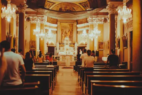 Wnętrze kościoła podczas mszy. Ludzie siedzą w ławkach, a przed ołtarzem trwa eucharystia.