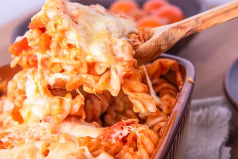 Un cucchiaio solleva una porzione generosa di pasta alla sorrentina, filante e dorata, da una pirofila marrone.