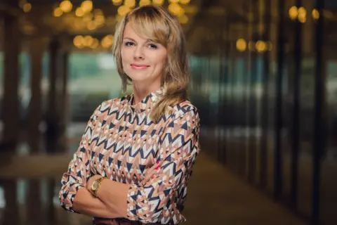 Portret uśmiechniętej kobiety w kolorowej bluzce, z rękami skrzyżowanymi na piersi.