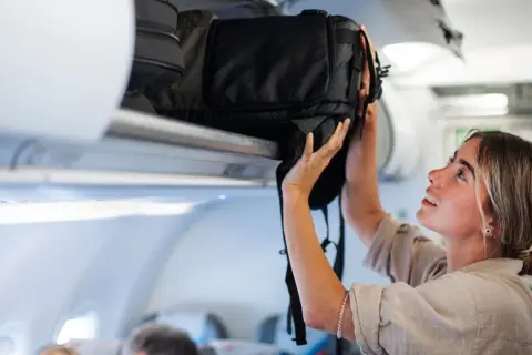 Junge Frau verstaut ihren großen Backpacking Rucksack im Gepäckfach eines Flugzeugs.