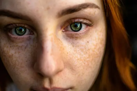Close-up texture photography of a young woman's face, highlighting her green eyes and the intricate pattern of her freckles.