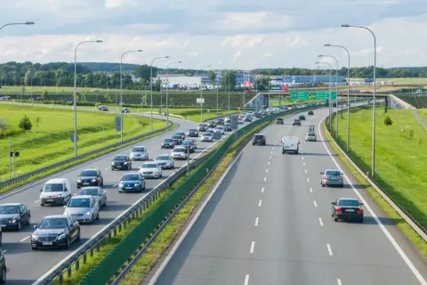Autostrada z ruchem samochodowym w słoneczny dzień, z zielonymi poboczami i drzewami w tle.