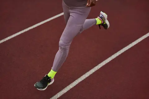 Une athlète court sur une piste, portant une chaussure de marche longue distance et des chaussettes fluo.