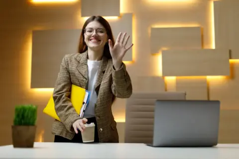 Uśmiechnięta kobieta w okularach, z teczkami i kawą, macha ręką. To jej dzień próbny w pracy bez umowy, pełen nadziei.