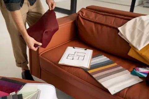 A person is choosing couch materials, holding a burgundy swatch next to a brown leather couch. Fabric samples and a floor plan are on the seat.