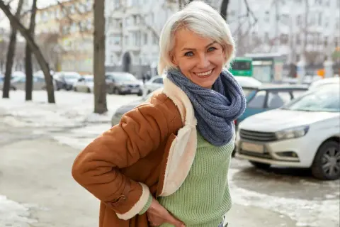 Uśmiechnięta kobieta w brązowym kożuchu i niebieskim szaliku stoi na tle zimowego miasta.