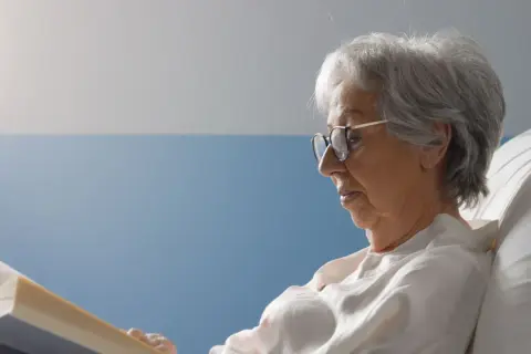 Une femme âgée, en **had et apa**, lit un livre confortablement installée dans un lit.