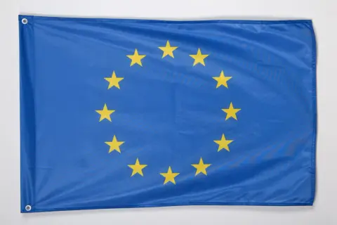 Niebieska flaga z dwunastoma złotymi gwiazdami ułożonymi w okrąg. To godło Unii Europejskiej, symbol jedności i solidarności.