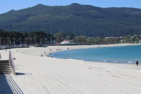 Amplia playa de arena blanca en la playa de Samil, con aguas turquesas y una montaña verde al fondo.