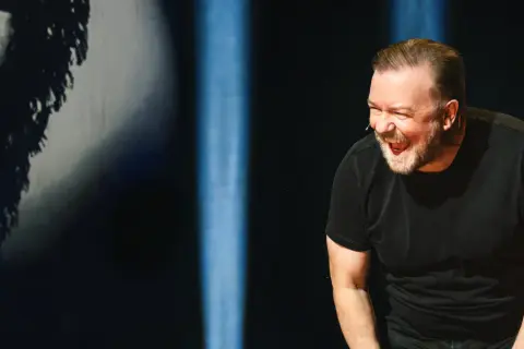 Ricky Gervais w swoim stand upie śmieje się do rozpuku, w czarnym t-shircie, na tle niebieskich świateł.