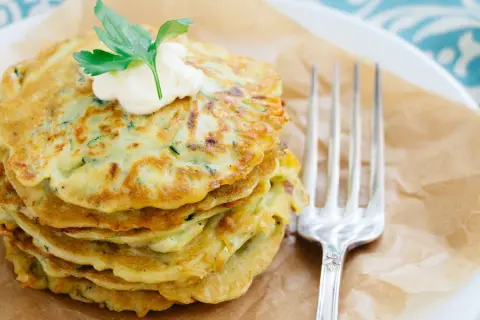 Ein Stapel goldbrauner Zucchini-Pancakes, garniert mit einem Klecks Sour Cream und Petersilie, daneben eine Gabel.