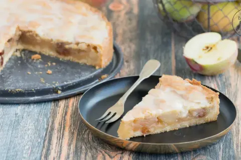 Ein Stück saftiger Apfelkuchen mit süßer Glasur und Mandelsplittern, daneben ein halber Apfel und ein Korb voller Äpfel.